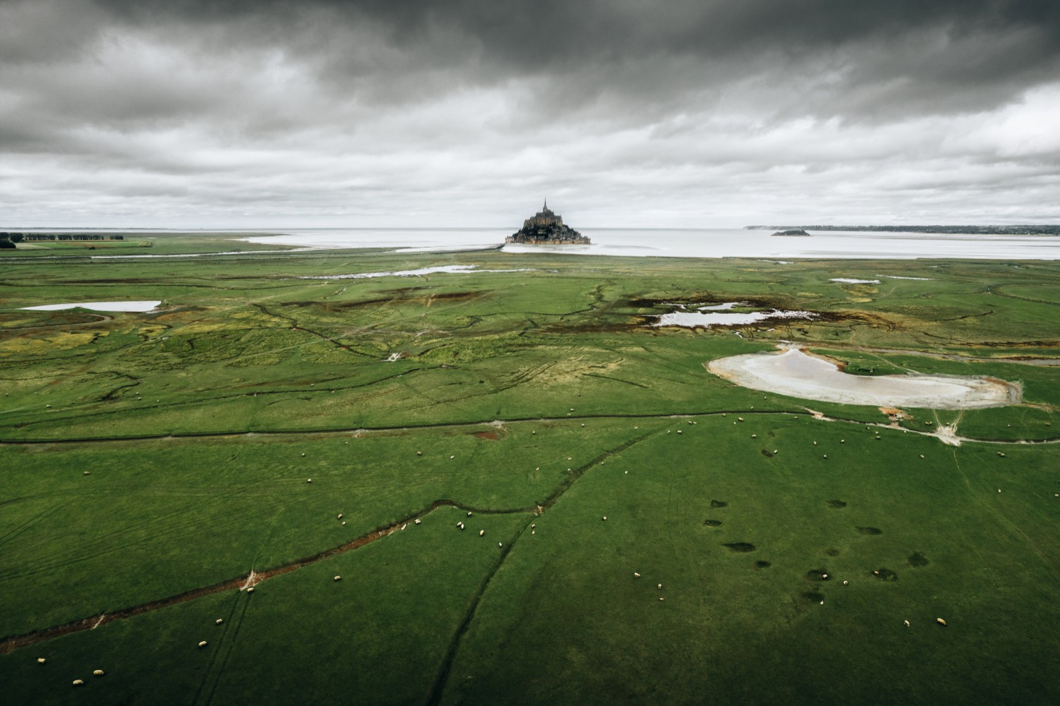 Prés salés et chenaux devant le Mont-Saint-Michel sous un ciel nuageux