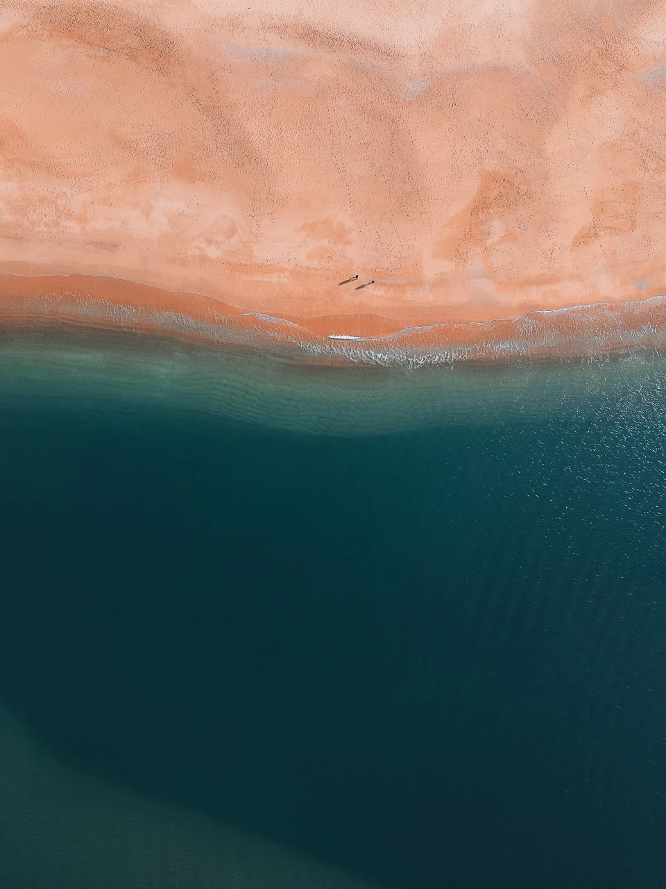 Plage rose et mer bleu sombre vues à la verticale, avec deux silhouettes sur le sable